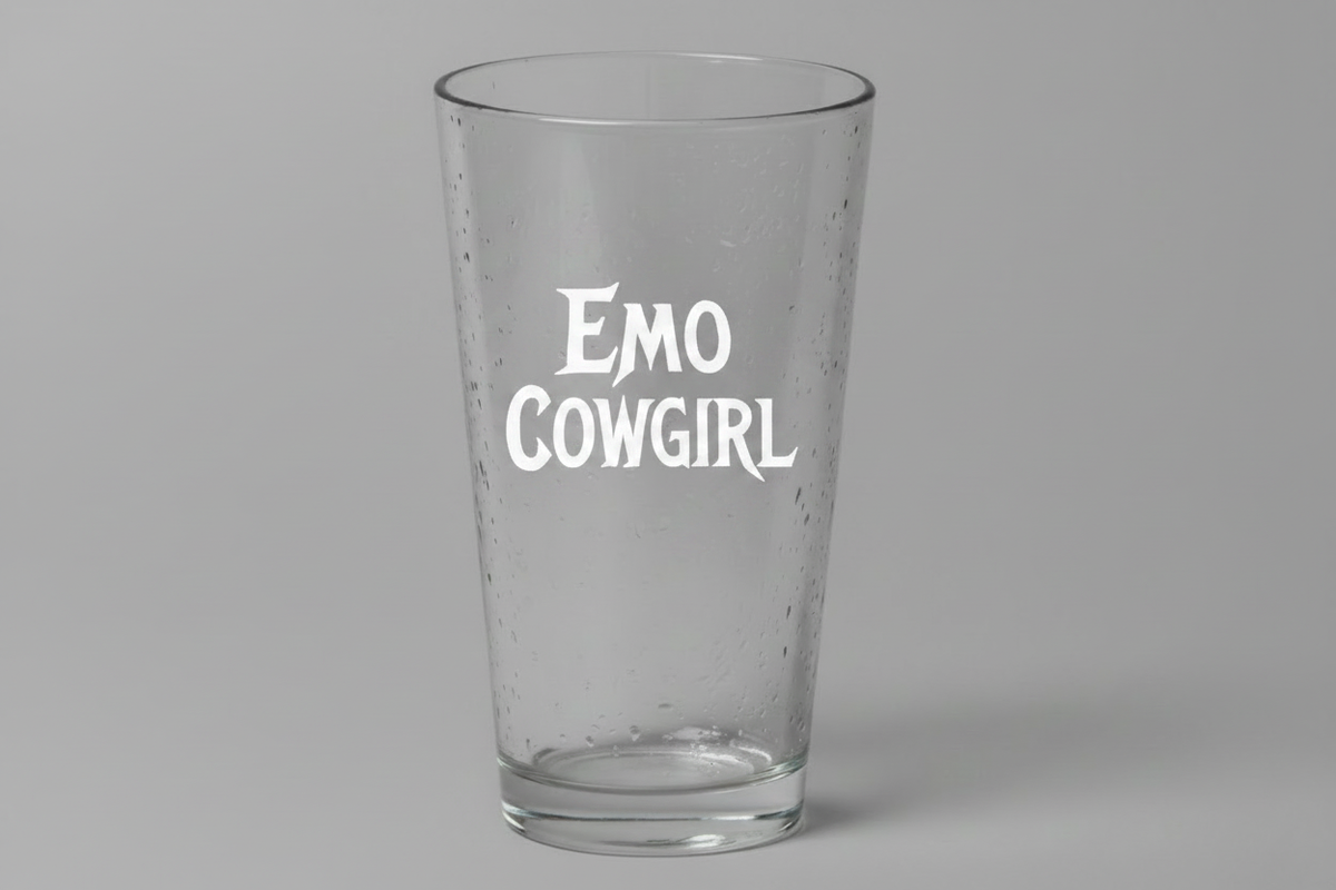 A clear pint glass with the words 'EMO COWGIRL' printed in white stylized text on the front, set against a plain gray background.