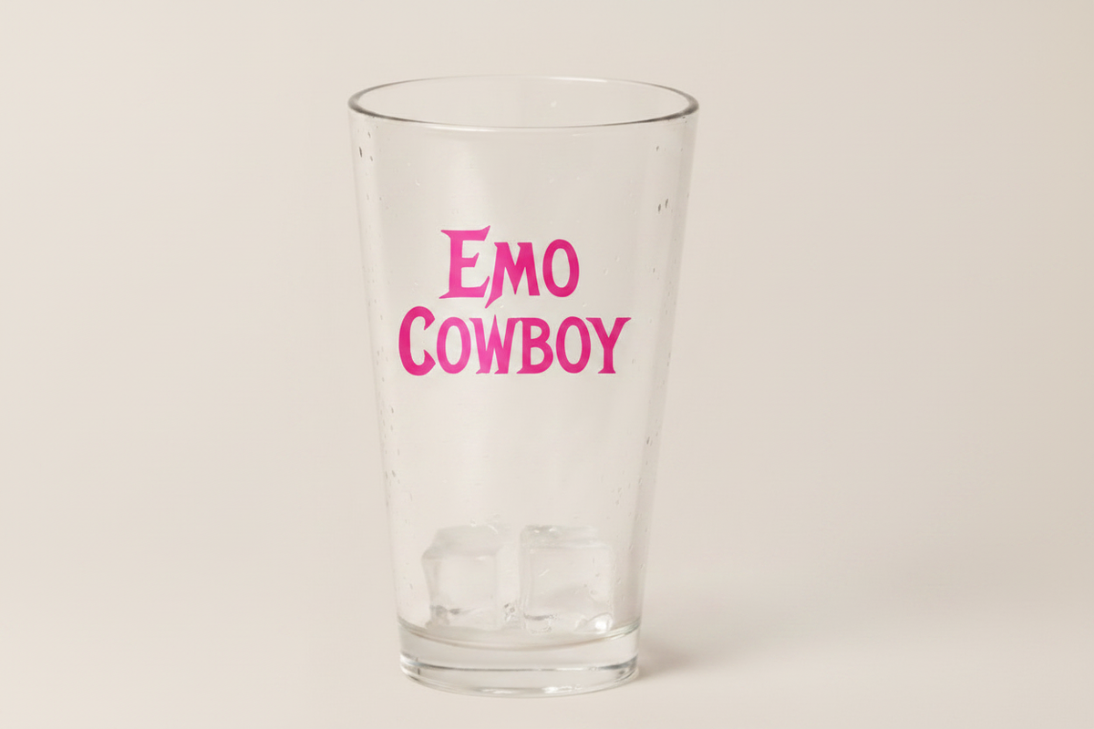 A clear glass pint glass with the words 'EMO COWBOY' printed in bright pink lettering, shown on a plain light beige background.