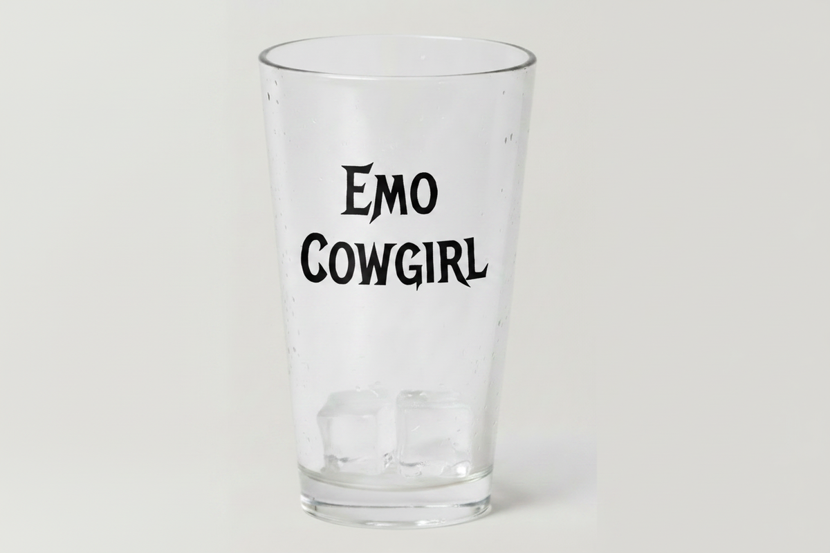 A clear glass pint cup with the words 'EMO COWGIRL' printed in black stylized text on the front, shown on a plain light gray background.