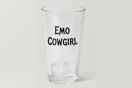 A clear glass pint cup with the words 'EMO COWGIRL' printed in black stylized text on the front, shown on a plain light gray background.