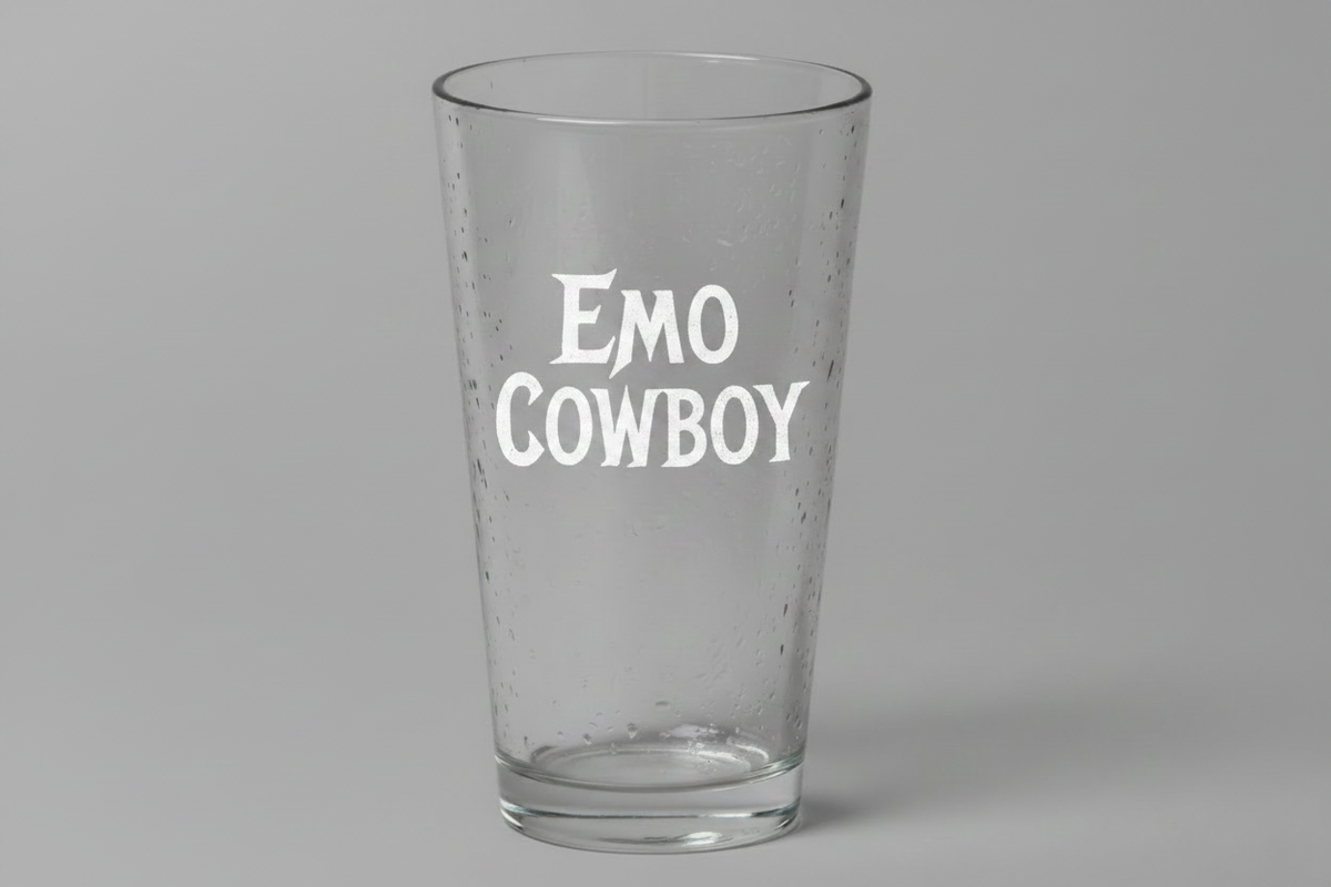 A clear glass pint glass with condensation droplets and the words 'EMO COWBOY' printed in white on the front, set against a solid gray background.