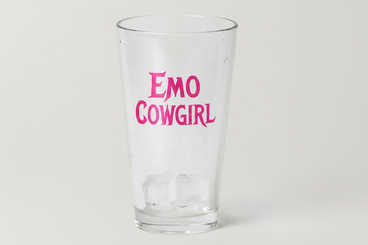 A clear glass pint cup with the words 'EMO COWGIRL' printed in bright pink stylized lettering, shown on a light gray background with ice cubes inside.