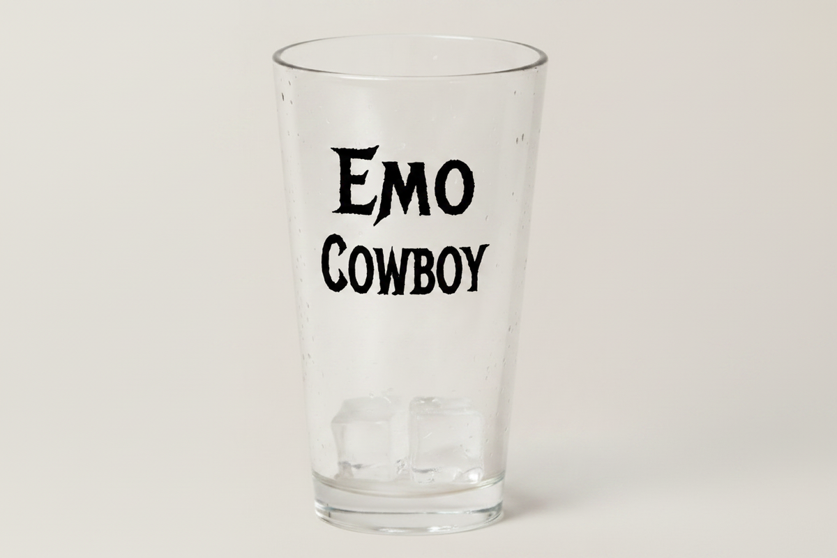 A clear glass pint glass with the words 'EMO COWBOY' printed in black gothic-style lettering on the front, shown on a light gray background.